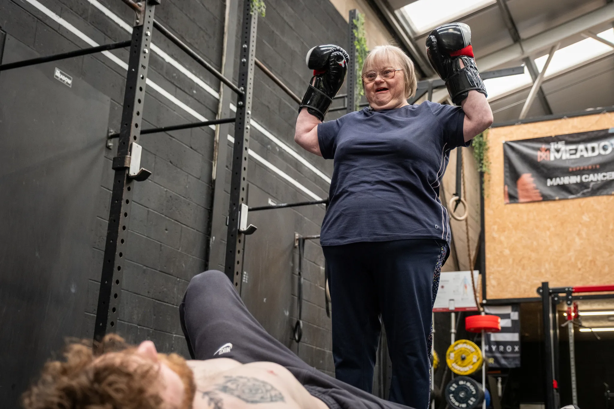A woman with Down's syndrome smiling triumphantly with both arms raised wearing black boxing gloves during a training session in a gym, with a trainer visible in the foreground and a Manx Cancer Care support banner visible in the background